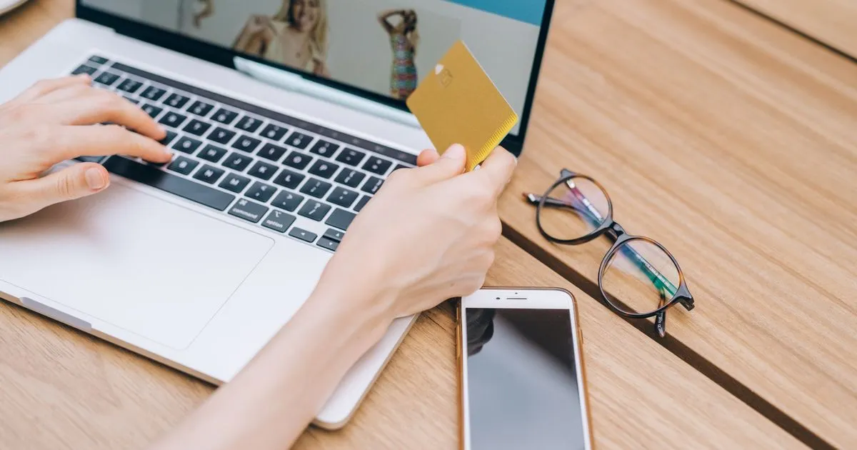 ノートパソコンでネットショップを見ながらクレジットカードを手にしている手元と、机に置かれたスマートフォン