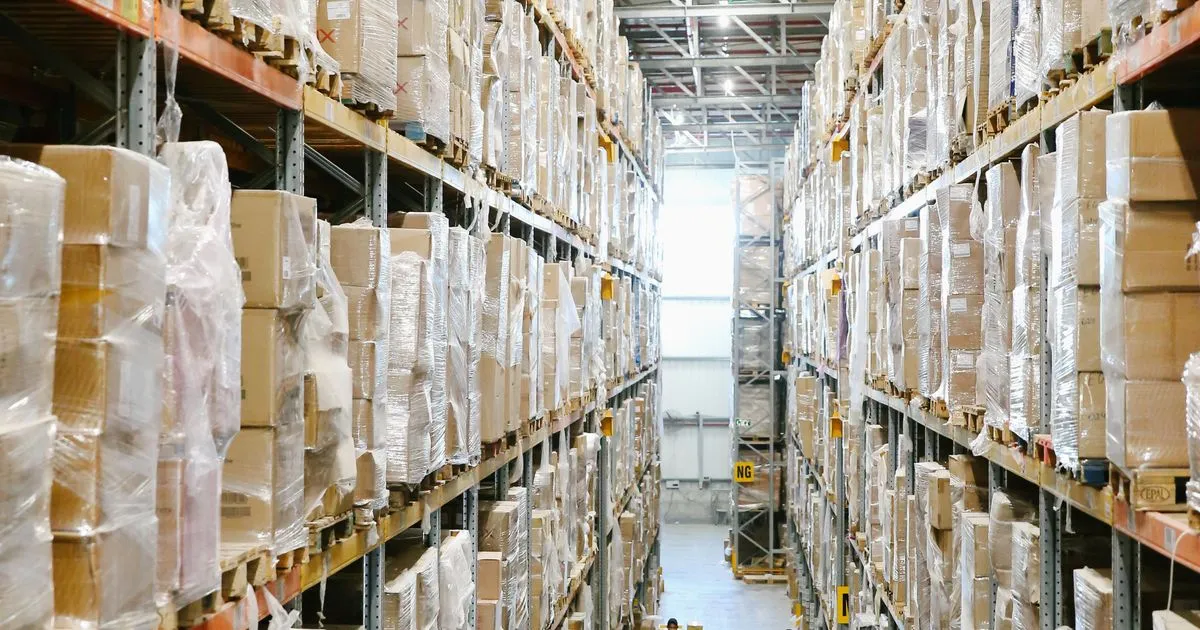 段ボールとストレッチフィルムで梱包された荷物が両側の棚に高く積まれた倉庫の通路
